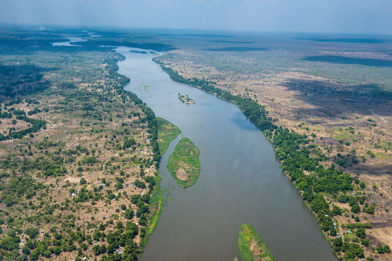 Imagem: Fotografia. Vista do alto de local com rio longo com água de cor escura, com parte com terras entre as água e vegetação. Nas bordas, à esquerda e à direita, local com solo bege, vegetação rasteira com árvores de folhas verdes e poucas moradias. Fim da imagem.