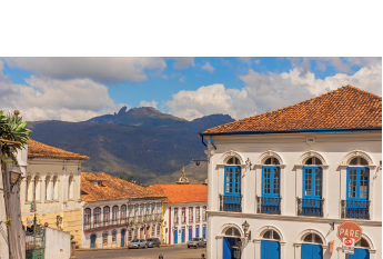 Imagem: Fotografia. Vista geral de local com rua cinza ao centro, com moto e piloto visto de costas. À esquerda e à direita, várias casas antigas de paredes de cor branca, com portas e janelas em cores variadas: amarela e azul e telhado de cor marrom-claro. Em segundo plano, morros altos com vegetação de cor verde-escuro. No alto, céu em azul-claro com nuvens brancas.  Fim da imagem.