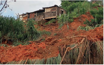 Imagem: Fotografia. Vista geral, de baixo para cima de lugar com vegetação, solo de cor marrom-escuro, com terra deslizada. Acima, árvores com folhas verdes e à esquerda, casas próximas umas das outras, com telhado na parte superior. No alto, céu em azul-claro com nuvens brancas.  Fim da imagem.