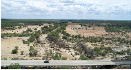 Imagem: Fotografia. Vista do alto de local com solo de cor bege, árvores com folhas verdes. À frente, estrada de cor cinza-claro e caminhão passando à esquerda. No alto, céu em azul-claro e nuvens brancas.  Fim da imagem.