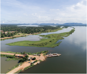 Imagem: Fotografia. Vista do alto de local com rio grande de água escura. À esquerda, local com vegetação de cor verde. À frente, solo de cor bege-claro, com extensão fina e contornos com verde. Perto do local embarcação de cor branca. No céu, azul-claro e nuvens brancas.  Fim da imagem.