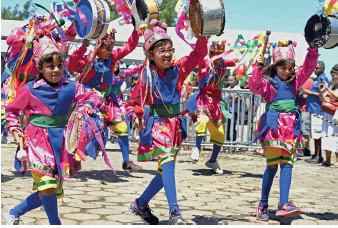 Imagem: Fotografia. Em um local com rua de cor cinza, com três crianças carregando um pequeno tambor na mão direita. Eles estão vestidos com roupa coloridas em rosa, detalhes e meiões em azul e bermuda até os joelhos em amarelo. Sobre a cabeça, gorro de cor rosa. Mais ao fundo, outras crianças vestidas como as outras. No alto, céu em azul-claro.  Fim da imagem.