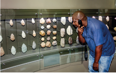 Imagem: Fotografia. Um homem agachado de frente para local com exposição de pedras na vertical sobre uma parede. No local há pedras pequenas, tamanho médio e grande com marrom, bege e cinza. O homem tem pele negra, careca, agachado, com camiseta em azul e calça jeans, com um aparelho preto perto do ouvido. Fim da imagem.