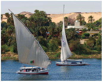 Imagem: Fotografia. Vista geral de local com rio de água escura, com dois barcos pequenos de cor branca, com vela alta em branco. Ao fundo, vegetação de cor verde, árvores e vista parcial de estabelecimentos de paredes claras. No alto, céu azul-claro sem nuvens.  Fim da imagem.