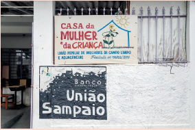 Imagem: Fotografia. Uma parede de cor branca, com ilustração à esquerda em cinza-escuro com texto em branco: Banco união Sampaio. No alto, grade de ferro na vertical com lanças na parte superior, placa de cor bege-claro, com texto em vermelho: Casa da Mulher e da criança. Na parte inferior em azul-claro: União popular de mulheres de campo limpo e adjacências. Fundada em 08/03/1987. À esquerda, vista parcial de local com mesas e cadeiras.  Fim da imagem.