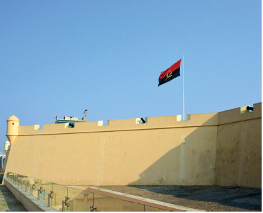 Imagem: Fotografia. Uma fortaleza de paredes de cor bege-claro, com pequena torre à esquerda e ao centro, uma bandeira hasteada de cor vermelha e preta. No alto, céu azul-claro.  Fim da imagem.