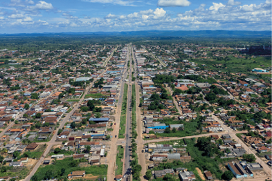 Imagem: Fotografia. Vista geral de alto, com rodovias de cor cinza ao centro. À esquerda e à direita, grama de cor verde, com quadras com casas e árvores de folhas em verde-escuro. Mais ao fundo, morros escuros. No alto, céu em azul-claro e nuvens brancas.  Fim da imagem.