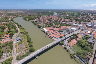 Imagem: Fotografia. Vista geral de alto de local com rio de cor verde ao centro, longo com uma ponte acima de cor cinza com automóveis passando. Nas bordas do rio à esquerda, árvores de folhas verdes e à direita, cidade com casas com telhados de cor marrom, entre árvores. No alto, céu de cor azul-claro com poucas nuvens brancas.  Fim da imagem.