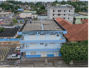 Imagem: Fotografia. Vista do alto de local com prédios de cor branca e partes em azul-claro, com três andares. No alto, título: Prefeitura de Cruzeiro do Sul. À esquerda, carros estacionados. À direita, casa grande, com telhado marrom e perto, vegetação de cor verde. Mais ao fundo, à direita, um prédio não terminado de cor cinza-claro. Em segundo plano, ruas, casas entre árvores de folhas verdes.  Fim da imagem.