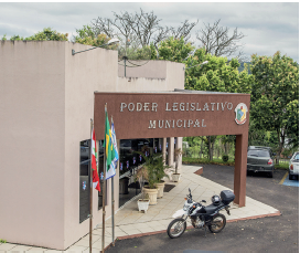 Imagem: Fotografia. Vista do alto de local com paredes de cor bege, entrada de cor marrom e na parte inferior, parede com vidros escuros. De frente da entrada, uma moto estacionada de cor preta. No alto do prédio, está escrito: PODER LEGISLATIVO MUNICIPAL. À esquerda, bandeiras hasteadas. Em segundo plano, carros estacionados e árvores com folhas verdes. No alto, céu nebuloso.  Fim da imagem.