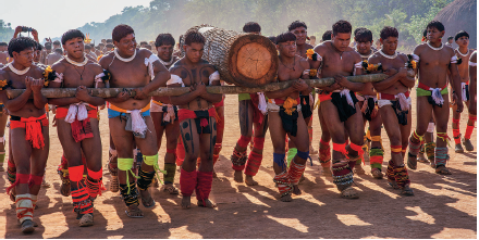 Imagem: Fotografia. Dezenas de homens em pé, morenos, de cabelos escuros, com tecido nas pernas e cintura de cor laranja, branca e partes em branco. De frente deles, um tronco na horizontal de madeira. Ao centro, entre eles, um tronco grande de cor marrom. Eles estão em solo de cor bege. Em segundo plano, morros e árvores de folhas verdes. Fim da imagem.