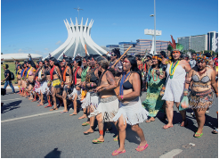 Imagem: Fotografia. Sobre rua de cor cinza com dezenas mulheres manifestando, uma ao lado da outra, caminhando em fileiras, para à esquerda. Ela tem pele morena, cabelos escuros, colares coloridos e par de chinelos. Ao fundo, algumas pessoas estão com uma coroa de penas coloridas. Em segundo plano, prédio com design para a vertical, com hastes finas. No alto, céu em azul-claro sem nuvens. Fim da imagem.