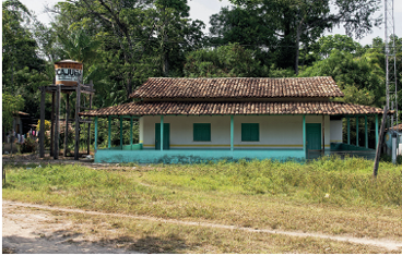 Imagem: Fotografia. Vista de local com grama de cor verde e mais ao fundo, escola com o formato de casa de paredes de cor branca com telhados de cor marrom, janelas e paredes de cor verde. Em segundo plano, árvores de folhas verdes. No alto, céu em azul-claro. Fim da imagem.