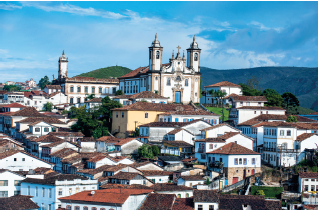 Imagem: Fotografia. Vista geral de local com casas antigas de paredes de cor branca, com telhado de cor marrom, triangular. Mais ao fundo, uma igreja de paredes de cor branca com detalhes em detalhes de cor amarela, com duas torres em cada ponta. Mais ao fundo, outra igreja de paredes brancas, com uma grande torre na vertical. À direita, morros de cor verde. No alto, céu em azul-claro com nuvens brancas.  Fim da imagem.