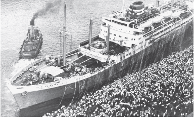 Imagem: Fotografia em preto e branco. Vista do alto de uma grande embarcação com algumas hastes na vertical, com pessoas dentro. À direita, centenas de pessoas e à esquerda, oceano com embarcação pequena por onde sai fumaça para o alto.  Fim da imagem.