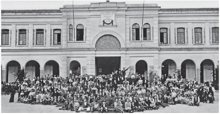 Imagem: Fotografia em preto e branco. À frente, centenas de pessoas de frente, homens de paletó e mulheres com saias longas e blusa de mangas compridas. Em segundo plano, estabelecimentos na horizontal de paredes claras, com dois andares com arcos e acima, na parte superior, janelas compridas na vertical. Mais ao centro, forma circular.  Fim da imagem.