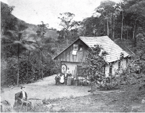 Imagem: Fotografia em preto e branco. Vista do alto de casa de madeira, com telhado triangular no alto. Ao redor, vegetação. Em primeiro plano, sentado, de terno e chapéu preto e camiseta branca por dentro. De frente para casa, dois homens em pé de terno, mulheres de blusas de mangas compridas e saia longa. Em segundo plano, densa vegetação.  Fim da imagem.