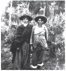 Imagem: Fotografia em preto. Dois homens vistos de frente, vestes longas escuras, com chapéu redondo na cabeça, com barba branca. À direita, uma mulher de pele morena com cabelos escuros com chapéu redondo, com blusa de mangas compridas clara e calça escura. Em segundo plano, vegetação.  Fim da imagem.