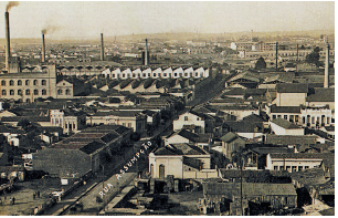 Imagem: Fotografia em tons de sépia. Vista do alto de cidade com uma rua central, com casas de paredes claras e telhado na parte superior. Ao fundo, à esquerda, usinas com grandes paredes, inúmeras janelas e torres de chaminés por onde sai fumaça para o céu.  Fim da imagem.