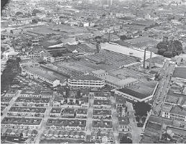 Imagem: Fotografia em preto e branco. Vista do alto de local com ruas, quadras com casarões e ao centro, muitos galpões com paredes claras e telhado escuro. Em algumas partes, árvores e mais ao fundo, outras casas.  Fim da imagem.