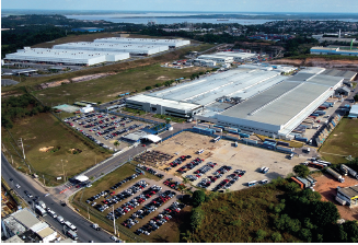 Imagem: Fotografia. Vista do alto de local com vários galpões grandes de cor cinza-claro, com muitos caminhões em volta e à esquerda, centenas de carros estacionados. Perto dos galpões, área com vegetação e árvores de folhas verdes e estrada à esquerda, por onde passam automóveis. Em segundo plano, outros galpões gigantes de cor branca. Em terceiro plano, área arborizada e rio.  Fim da imagem.