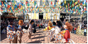 Imagem: Fotografia. Vista geral de local com bonecos grandes dançando. À esquerda, bonecos vestidos de homens: um de pele clara, cabelos pretos, outro ruivo e ao lado dele, um boneco negro de cabelos pretos. Todos usam uma camisa azul com estampa em laranja, de mangas compridas e calça em bege. À direita, outras bonecas dançando de blusa de mangas compridas e saia longa de cor azul com estampas em laranja. Uma delas tem cabelos loiros, outra tem cabelos pretos e outra tem cabelos castanhos. No alto, linha finas com bandeiras coloridas penduradas. Mais ao fundo, prédio de paredes amarelas com partes em branco;  Fim da imagem.