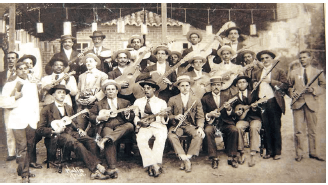 Imagem: Fotografia em tons de sépia. Dezenas de homens sentados um ao lado do outro, com chapéu redondo, com terno e sapatos de couro. Alguns estão com terno claro, outros escuros, segurando nas mãos instrumentos musicais: flautas finas e cavaquinho à frente e ao fundo, alguns homens com violão, em pé. Mais ao fundo, na parte superior, luzes penduradas.  Fim da imagem.