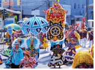 Imagem: Fotografia. Em uma rua, pessoas fantasiadas de roupas em azul e detalhes em preto e amarelo. À direita, criança com roupa em vermelho e detalhes em branco. Ao centro, pessoa morena com vestido grande preto e detalhes em amarelo e branco. Mais ao fundo, outra mulher de vestido roxo, detalhes em amarelo e partes em laranja. Em segundo plano, um tecido grande na vertical de cor vermelha com detalhes em amarelo, partes em azul-escuro e texto em branco: Estrela- Brilhante. Mais ao fundo, outros dançarinos e pessoas fantasiadas. Fim da imagem.