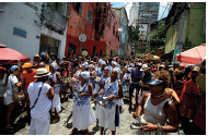 Imagem: Fotografia. Rua de cor cinza, com dezenas de pessoas negras com vestidos de cor branca, colares, com lençol na cabeça, em azul-claro. À esquerda e à direita, casas de paredes em branco , vermelho e verde. Sobre as paredes, antenas de televisão redonda e ao fundo, prédio de cor cinza. No céu, azul-claro com nuvens brancas.  Fim da imagem.