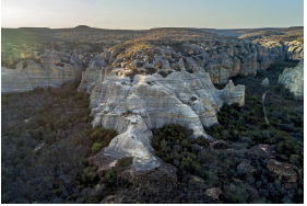 Imagem: Fotografia. Vista do alto de local com rochas grandes de cor bege, disformes, com pequenos buracos na parte superior. Na parte inferior das rochas, vegetações de cor verde-escuro. O céu é de cor azul-claro sem nuvens.  Fim da imagem.