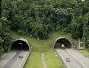 Imagem: Fotografia. Vista geral do alto de local com vegetação verde e árvores com folhas verdes. À esquerda e à direita, rua cinza por onde passam carros e ao fundo, entrada arredondado para túnel.  Fim da imagem.