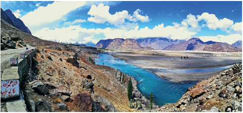 Imagem: Fotografia. Vista do alto de local com morros e montanhas de cor marrom e ao centro, rio com água de cor azul. Em segundo plano, montanhas e no alto, céu de cor azul com nuvens.  Fim da imagem.