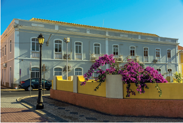 Imagem: Fotografia. Vista geral de local com muro de cor amarela, com flores de pétalas de cor roxa e à esquerda, calçada com luminária de rua em cinza-escuro e luz em amarelo. Mais ao fundo, um casarão de paredes de cor azul-claro e detalhes em branco nas janelas e nas colunas. No alto, céu em azul-claro. Fim da imagem.