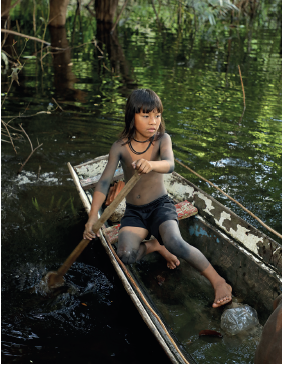 Imagem: Fotografia. Dentro de uma canoa fina sobre rio de cor verde, com menina indígena sentada. Ela tem pele morena, com cabelos pretos com franja, manchas de cor cinza no corpo, com bermuda em preto, sem sapatos e segurando nas mãos, um remo de cor marrom. Ela olha para à direita, com colar preto no pescoço. Na água, vegetação dentro.  Fim da imagem.