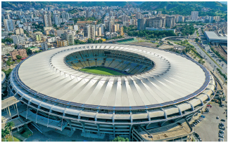 Imagem: Fotografia. Vista do alto de local arredondado de cor cinza, com abertura ao centro e abaixo, vê-se grama verde e ao redor, as arquibancadas. Em segundo plano, cidade vista de prédios de tamanhos diferentes.  Fim da imagem.