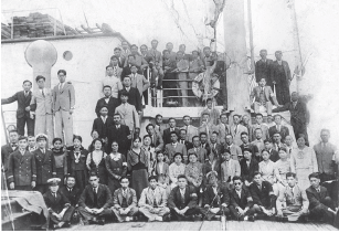 Imagem: Fotografia. Dezenas de homens posando para foto dentro de uma embarcação. Ao centro, eles estão sentados de pernas cruzadas, vestidos de terno e gravata, de pele clara e cabelos escuros. Outros estão em pé e à esquerda, pessoas em pé sobre escada e no andar de cima. À direita, uma haste na vertical.  Fim da imagem.