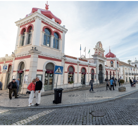 Imagem: Fotografia. À frente, rua de cor cinza, calçada com pessoas caminhando. À esquerda, uma mulher e um homem um ao lado do outro, perto de uma placa azul indicando faixa de pedestre. A mulher de tem morena, cabelos grisalhos, blusa de mangas compridas rosa, calça e sapatos brancos. O homem tem pele morena, cabelos grisalhos, com blusa de mangas compridas e sapatos pretos e calça branca. Mais ao fundo, prédio de paredes de cor bege com dois andares, com janelas compridas e parte superior arredondada. No alto céu de cor azul e nuvens esparsas.  Fim da imagem.