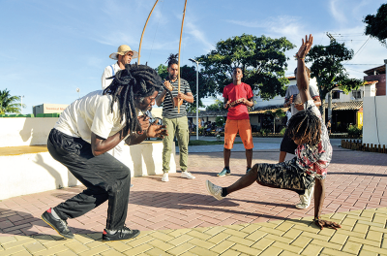 Imagem: Fotografia. Duas pessoas em uma calçada marrom praticando capoeira. À esquerda, um homem negro agachado com camiseta branca, calça e sapatos pretos, batendo palmas e cabelos escuros. À direita, menino de pele negra, cabelos escuros, com camiseta em branco e estampas em preto e vermelho, bermuda preta e sapatos brancos, se equilibrando com braço direito na vertical e outro esticado para o alto. Mais ao fundo, três pessoas em pé morenas, segurando nas mãos instrumentos, um homem visto parcialmente com camiseta branca, chapéu bege, com berimbau. À direita, um homem com camiseta com listras em branco e preto na horizontal e calça bege, sapatos brancos, berimbau. Ao ado, homem moreno, com camiseta vermelha e bermuda laranja.  Fim da imagem.