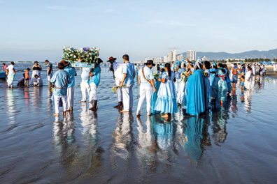 Imagem: Fotografia. Dezenas de pessoas sobre areia com água do mar em pé com roupas em branco e azul-claro, enfileirado. Há mulheres de vestidos longos, em azul-claro e outras em branco e homens com camiseta azul com calça branca. À esquerda, dois homens morenos, com chapéu marrom, visto de costas com objeto azul nos ombros com flores de pétalas brancas. Mais à esquerda, outras pessoas, crianças, uma canoa vista parcialmente, com ondas do mar. Em segundo plano, cidade e prédios de paredes brancas, com morros à direita. No alto, céu azul-claro.  Fim da imagem.