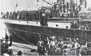 Imagem: Fotografia em preto e branco. À frente, várias pessoas em pé, com paletó, chapéu sobre a cabeça. Ao fundo, navio grande com centenas de pessoas de palito e chapéu na cabeça. Tem pessoas em pé perto do mastro.  Fim da imagem.