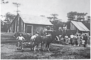 Imagem: Fotografia em preto e branco. Duas casas de madeiras com telhados triangulares. À frente, homens de blusa branca, calça escuro e mulheres com blusa e saia longa. Entre eles, duas crianças. À frente deles, cavalos e um homem de blusa clara e calça escura. Em segundo plano, árvores.  Fim da imagem.