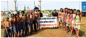 Imagem: Fotografia. Lugar com solo de cor marrom-claro, com dezenas de crianças. À esquerda, meninos indígenas de pele morenas, sem camiseta, com coroa de penas coloridas na cabeça, com bermuda longa e segurando nas mãos, lança fina na ponta. Ao centro, faixa de cor branca, texto em preto e vermelho. À direita, meninas umas perto das outras, de pele morena, cabelos longos escuros, usando blusa curta e saias coloridas em verde, vermelho, rosa. Sobre o pescoço delas, colares. No alto, céu azul-claro e nuvens brancas esparsas.  Fim da imagem.