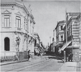 Imagem: Fotografia em preto e branco. Vista de local com rua ao centro e à esquerda e à direita, casarões grandes e antigos. Um à esquerda, com dois andares, com janelas grandes e finas. Mais ao fundo, casas similares.  Fim da imagem.