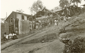 Imagem: Fotografia em tons de sépia. Vista de local com morro com vegetação rasteira e à esquerda, casas de paredes e muros de madeira, com telhado triangular. De frente das casas, pessoas descendo e mais abaixo, outras três sentadas junto com uma criança de blusa clara, e as mulheres de saia longa. Perto das casas, ao fundo, árvores de folhas verdes.  Fim da imagem.