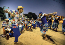 Imagem: Fotografia. Sobre local com solo de cor bege, arenoso, mulheres e homens, dois dançando. À esquerda, um homem de pele morena, cabelos pretos, com chapéu de palha sobre a cabeça, camiseta com estampas coloridas em azul e vermelho e calça azul, dançando e se equilibrando sobre uma perna só. À frente, à direita, mulher de pele morena, cabelos pretos, com lenço azul sobre a cabeça, blusa azul e com estampas na gola e saia longa estampada, com os braços abertos. À esquerda, dezenas de homens sentados, tocando instrumentos. Eles são negros, usam chapéu bege redondo sobre a cabeça, com camiseta estampada de cor azul e partes em vermelho e calça azul. Mais ao fundo, árvore de folhas verdes e céu em azul-claro, sem nuvens.  Fim da imagem.