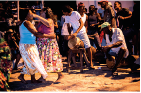 Imagem: Fotografia. Duas mulheres dançando à esquerda. Elas são negras, de cabelos escuros encaracolados, uma de frente para outra. À esquerda, mulher de regata branca, saia longa em branco e laranja e à frente, uma mulher de regata rosa e saia longa com flores. À direita, um homem negro agachado, sobre um tambor de cor bege, com boné verde, camisa branca e bermuda cinza. Ao lado, um moço de pele morena, cabelos escuros, com camiseta de cor branca, bermuda cinza, com as mãos sobre um tambor marrom sobre um cavalete. Mais ao fundo, outros homens negros dançando.  Fim da imagem.