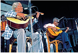 Imagem: Fotografia. De frente para um microfone, dois homens sentados tocando violão, sentados. À esquerda, um homem de pele morena, cabelos grisalhos, com camisa de mangas compridas xadrez em branco e azul, com calça cinza, tocando violão de cor bege. Mais à direita, homem moreno de cabelos escuros, com óculos de sol preto, camiseta de cor branca, com calça em azul, tocando violão de cor amarela e de pernas cruzadas.  Fim da imagem.