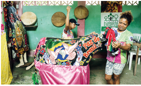 Imagem: Fotografia. Um boi com tecidos coloridos para à direita, com parte em preto, duas pombas brancas e bandeira do Brasil. No pescoço, texto em vermelho: Paz do Brasil. Sobre a cabeça, fitas de cor vermelha e tecido rosa na parte inferior. De frente para boi, mulher de pele negra, cabelos castanhos encaracolados para trás, com camiseta verde e rosa e bermuda em branco e cinza. Mais atrás, um homem visto parcialmente, com braço para o alto segurando uma linha. Ele tem pele morena, boné preto de camiseta branca com estampa. Mais ao fundo, parede de cor verde-claro, com objetos redondos marrons e à direita, faixa pendurada na vertical.  Fim da imagem.