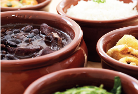 Imagem: Fotografia. Vista do alto de potes de barro redondos em marrom, com alimentos dentro. À esquerda, feijoada de cor marrom-escuro, à direita, vegetais em verde, outro com alimento bege e mais ao fundo, pote com arroz branco.  Fim da imagem.