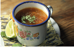 Imagem: Fotografia. Vista do alto de uma caneca de cor branca, com borda azul na parte superior e estampa de flores. Dentro da caneca, caldo de cor marrom com folhas verdes. Ao lado, à esquerda, pedaço de limão, debaixo para tecido sobre prato branco redondo.  Fim da imagem.